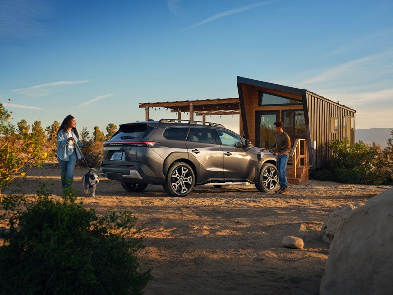 A static image of a blue 2026 Subaru Trailseeker parked on a grassy clearing in a rocky mountain landscape.
