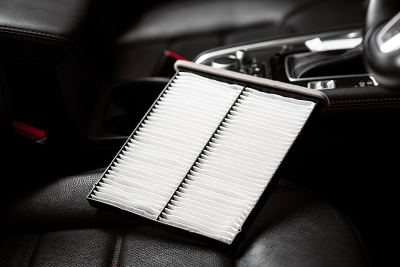 A new, rectangular white cabin air filter resting on a dark car seat with the car's interior visible in the background.