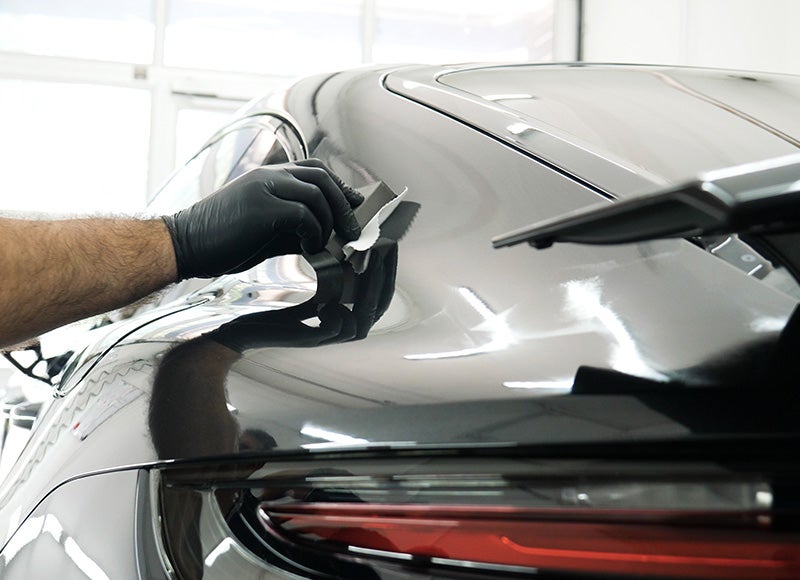 A gloved hand applies a ceramic coating with a sponge to a shiny black car's body.